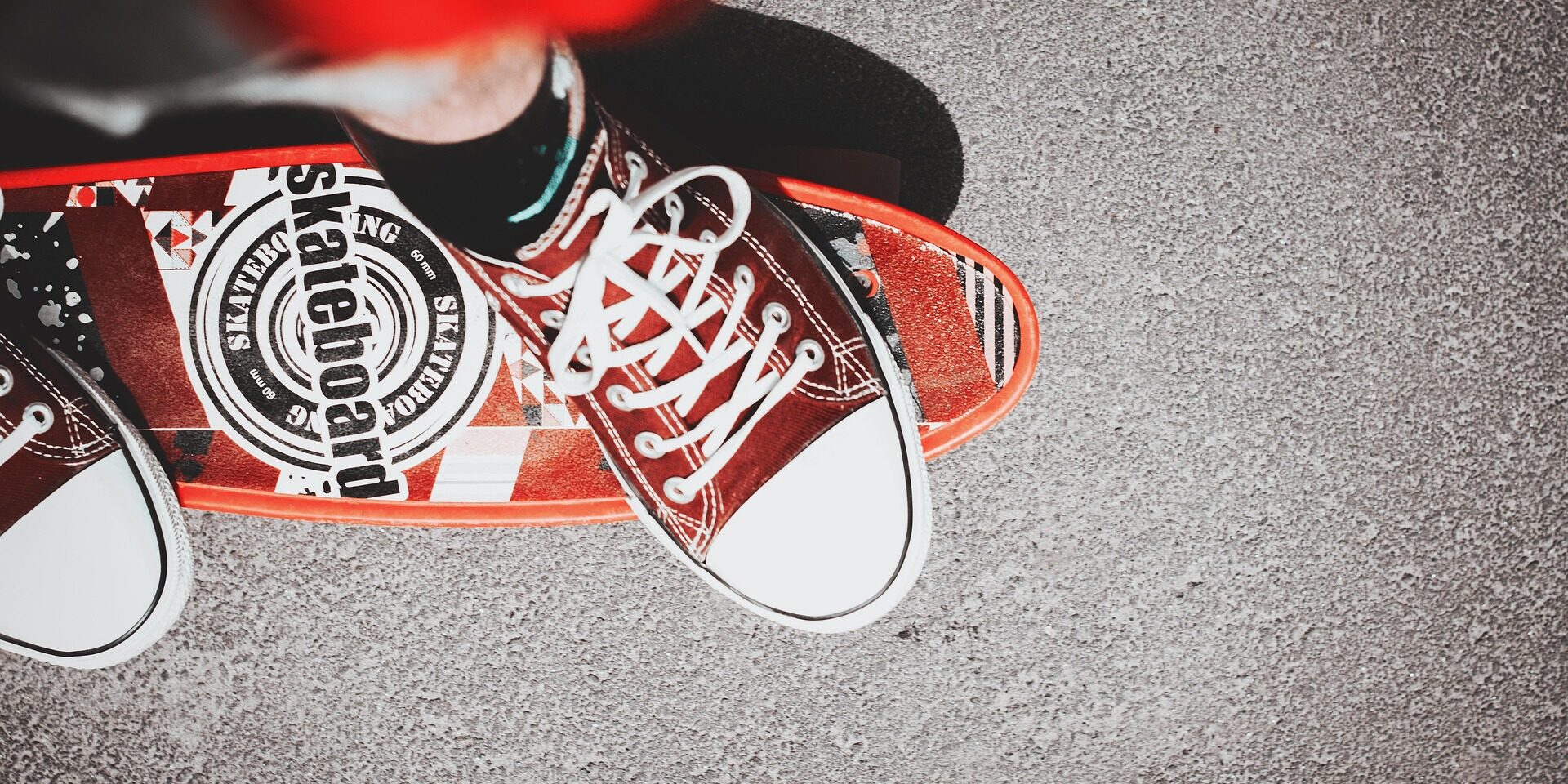 Red trainers standing on a red skateboard on tarmac, viewed from above.